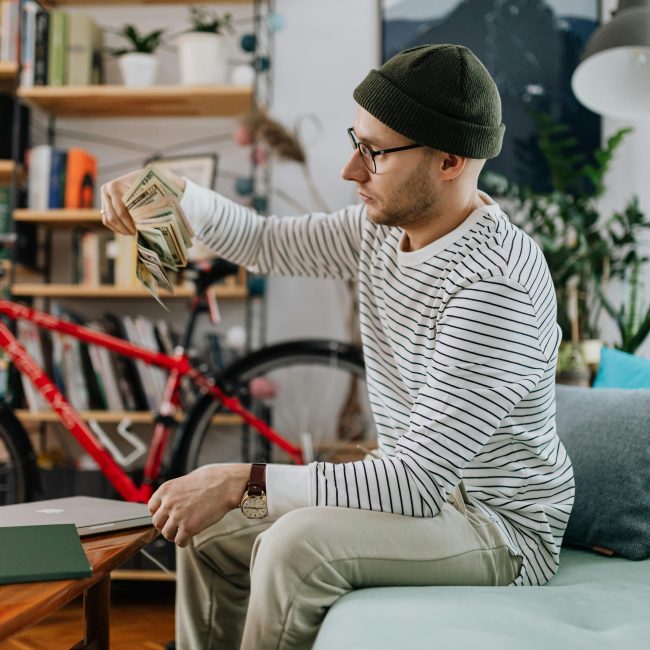Un homme en tenue décontractée comptant de l'argent assis sur un canapé dans un cadre domestique avec un vélo en arrière-plan.