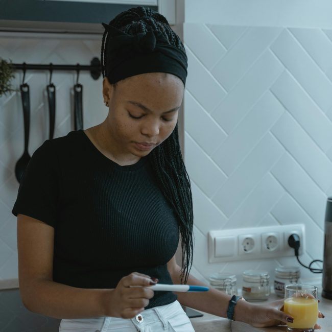 Une jeune femme dans une cuisine tenant un test de grossesse, plongée dans ses pensées.