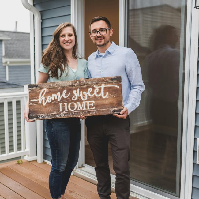 Couple souriant tenant une pancarte "Home Sweet Home" devant leur nouvelle maison