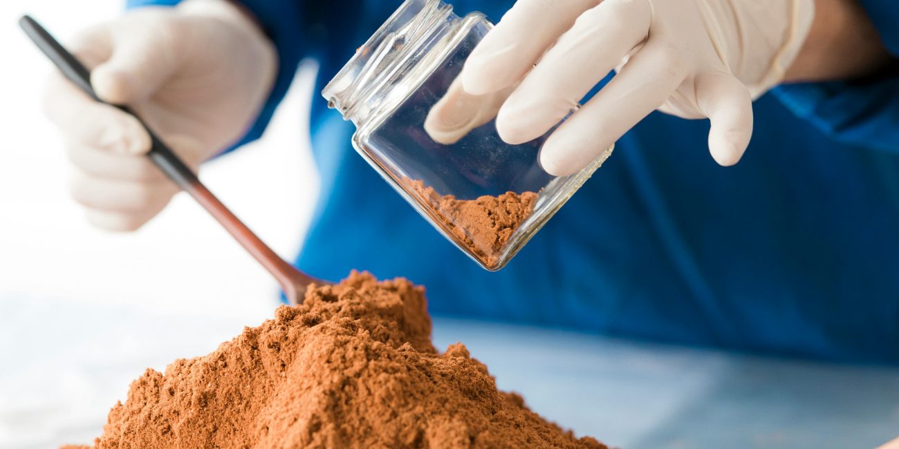 Un scientifique en tenue bleue analyse de la poudre de sol avec des gants et des cuillères en bois.