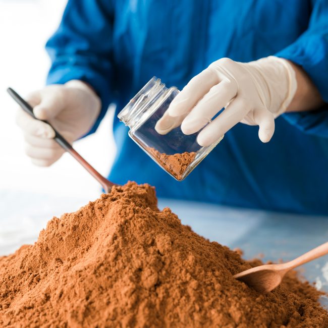 Un scientifique en tenue bleue analyse de la poudre de sol avec des gants et des cuillères en bois.