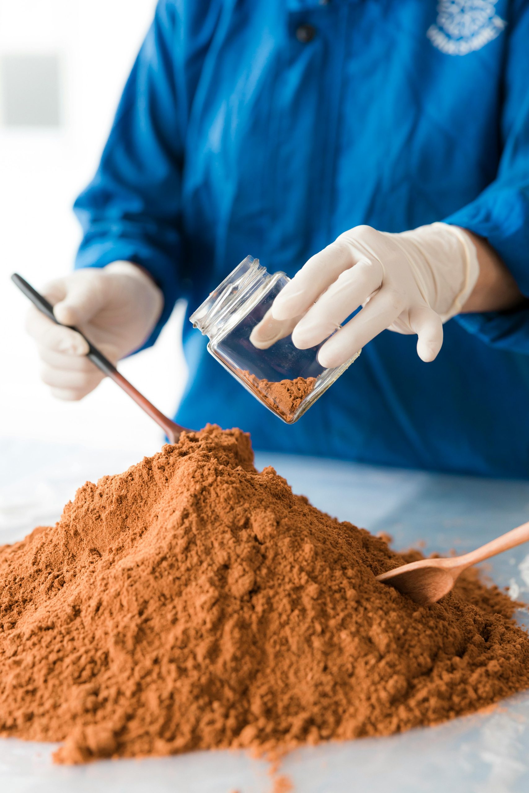 Un scientifique en tenue bleue analyse de la poudre de sol avec des gants et des cuillères en bois.