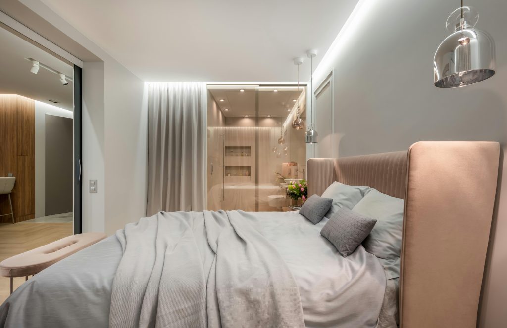 Intérieur d'une chambre moderne lumineuse avec des oreillers et une couverture sur le lit sous des lampes à côté d'un pouf près d'un mur vitré avec rideau