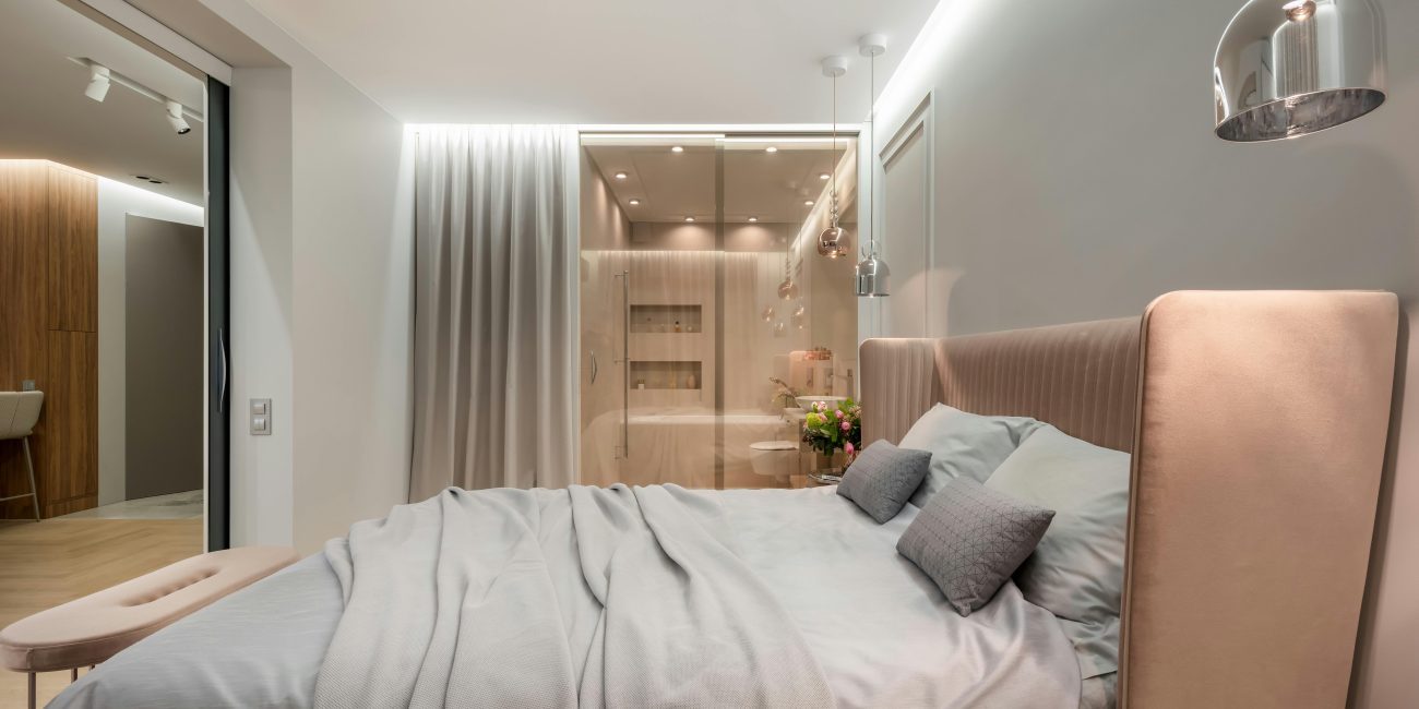 Intérieur d'une chambre moderne lumineuse avec des oreillers et une couverture sur le lit sous des lampes à côté d'un pouf près d'un mur vitré avec rideau