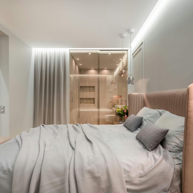 Intérieur d'une chambre moderne lumineuse avec des oreillers et une couverture sur le lit sous des lampes à côté d'un pouf près d'un mur vitré avec rideau