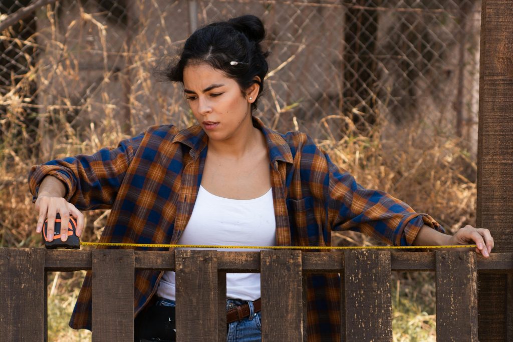 Focused woman using measuring tape on fence, showcasing empowerment in carpentry. Femme concentrée utilisant un mètre ruban sur une clôture, illustrant l'autonomisation en menuiserie.