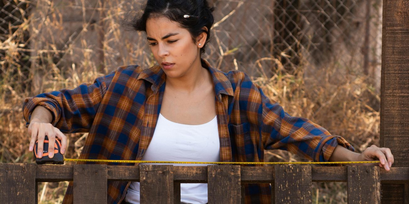 Focused woman using measuring tape on fence, showcasing empowerment in carpentry. Femme concentrée utilisant un mètre ruban sur une clôture, illustrant l'autonomisation en menuiserie.