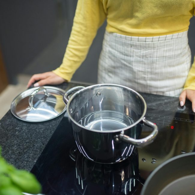 Une femme en pull jaune cuisinant sur une plaque à induction avec une casserole en inox.