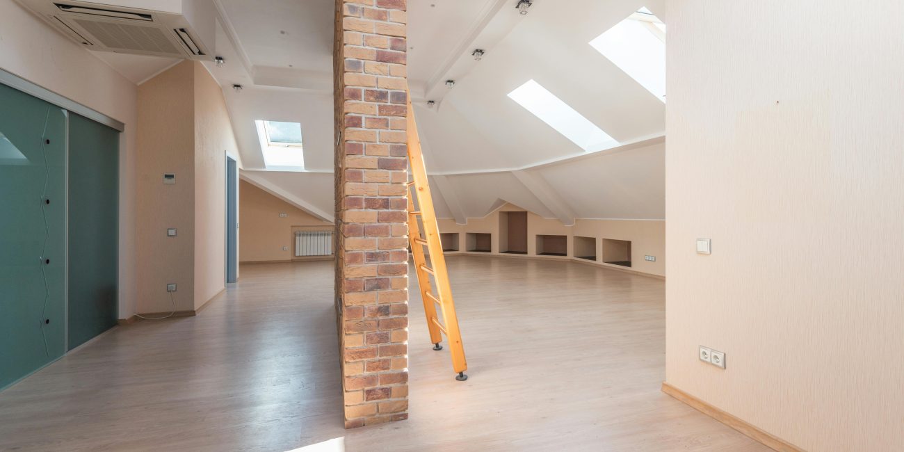Comble spacieux baigné de lumière naturelle grâce à des puits de lumière, avec un pilier en brique et une échelle en bois.