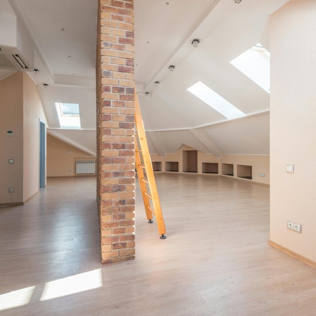Comble spacieux baigné de lumière naturelle grâce à des puits de lumière, avec un pilier en brique et une échelle en bois.