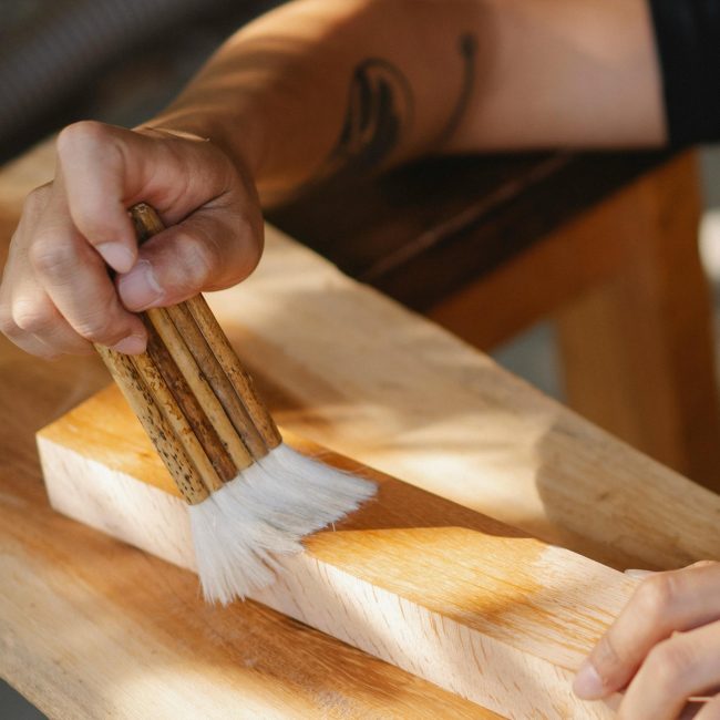 High angle of unrecognizable artisan using brush for varnishing wooden beam while working in professional workshop Vue de haut d'un artisan méconnaissable utilisant un pinceau pour vernir une poutre en bois tout en travaillant dans un atelier professionnel