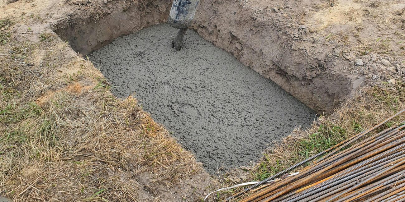 Concrete being poured into a foundation pit at a construction site in Poland, showing rebar and worker boots. Du béton versé dans une fosse de fondation sur un chantier de construction en Pologne, montrant les armatures et les bottes des ouvriers.