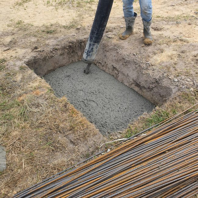 Concrete being poured into a foundation pit at a construction site in Poland, showing rebar and worker boots. Du béton versé dans une fosse de fondation sur un chantier de construction en Pologne, montrant les armatures et les bottes des ouvriers.