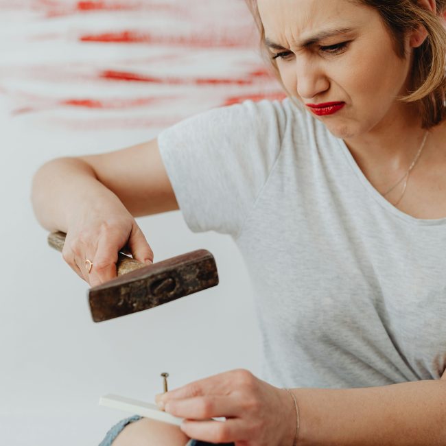 A woman struggling with a DIY project, exhibiting frustration with a hammer and nail. Une femme en difficulté avec un projet de bricolage, montrant de la frustration avec un marteau et un clou.