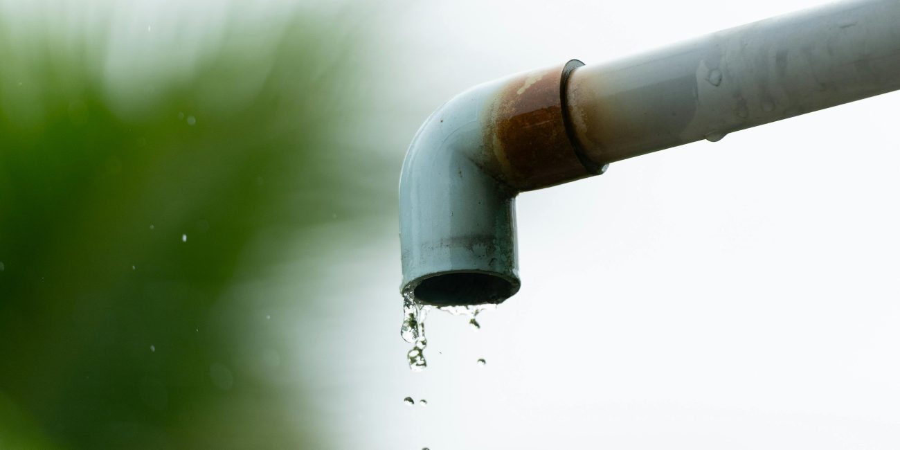 A close-up of water droplets falling from a rusty outdoor pipe against a blurred natural background. Gros plan de gouttes d'eau tombant d'un tuyau extérieur rouillé sur un fond naturel flou.