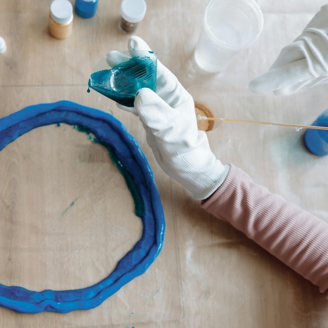 Close-up of hands crafting blue resin art indoors, showcasing artistic process. Gros plan sur des mains créant de l'art en résine bleue en intérieur, montrant le processus artistique.