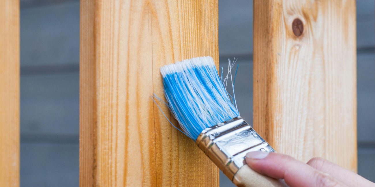 A hand painting a wooden fence plank with a blue paintbrush outdoors. Perfect for DIY and renovation themes. Une main peignant une planche de clôture en bois avec un pinceau bleu à l'extérieur. Parfait pour les thèmes de bricolage et de rénovation.