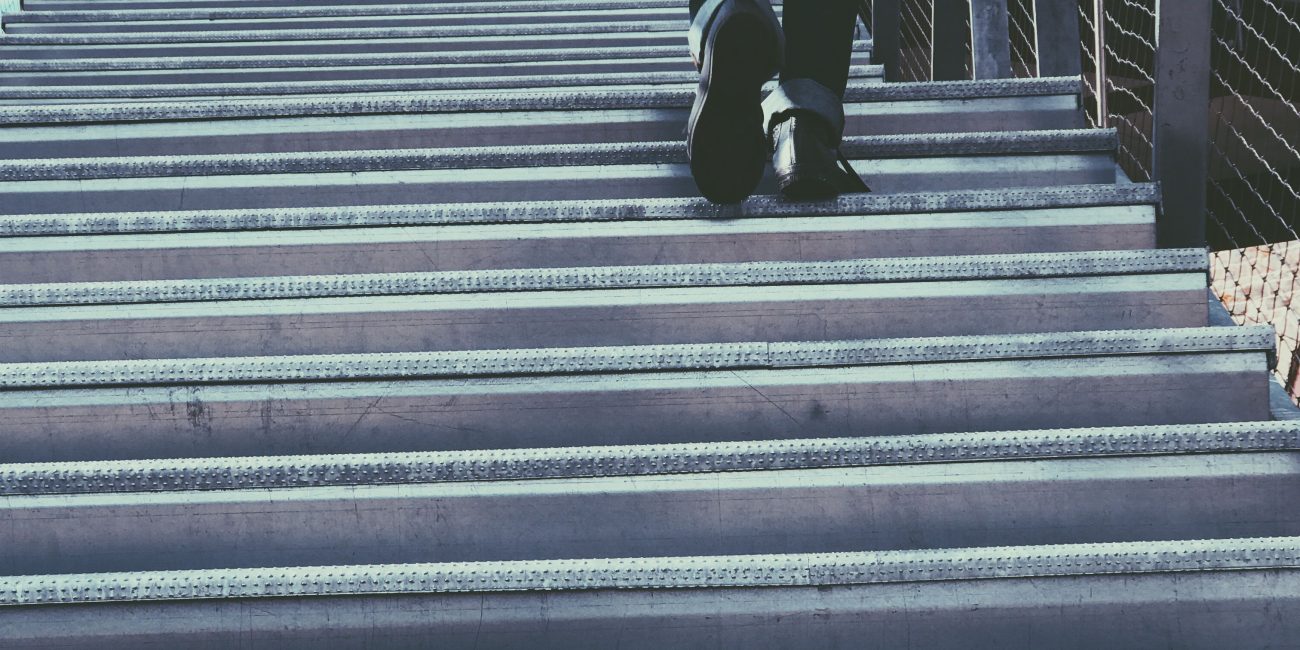 A person in casual wear ascends a metal staircase in an urban setting, suggesting travel or progress. Une personne en tenue décontractée monte un escalier métallique dans un cadre urbain, suggérant un voyage ou une progression.