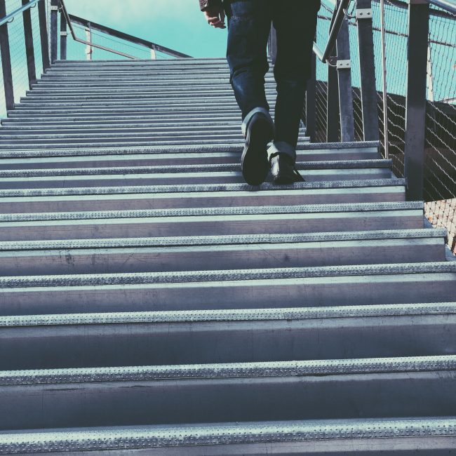 A person in casual wear ascends a metal staircase in an urban setting, suggesting travel or progress. Une personne en tenue décontractée monte un escalier métallique dans un cadre urbain, suggérant un voyage ou une progression.