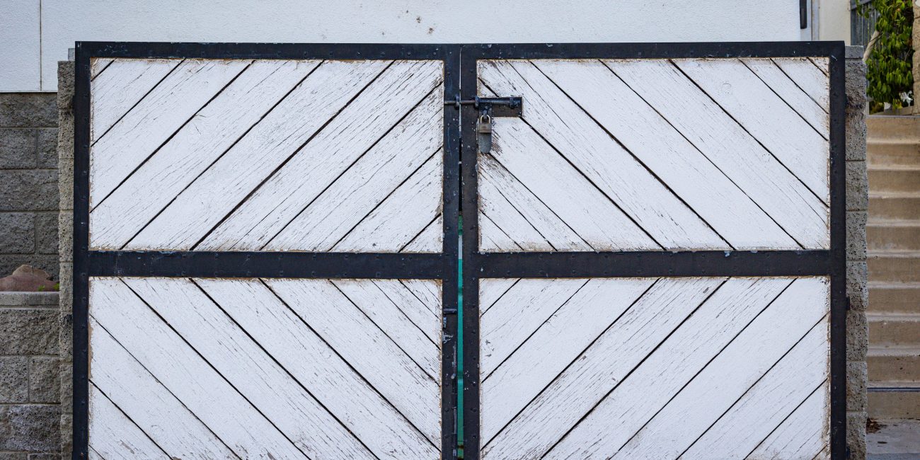 A rustic white wooden gate with diagonal panels and a black metal frame in a residential entrance. Un portail blanc rustique en bois avec des panneaux diagonaux et un cadre en métal noir à l'entrée d'une résidence.