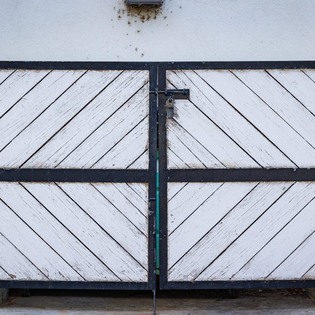 A rustic white wooden gate with diagonal panels and a black metal frame in a residential entrance. Un portail blanc rustique en bois avec des panneaux diagonaux et un cadre en métal noir à l'entrée d'une résidence.
