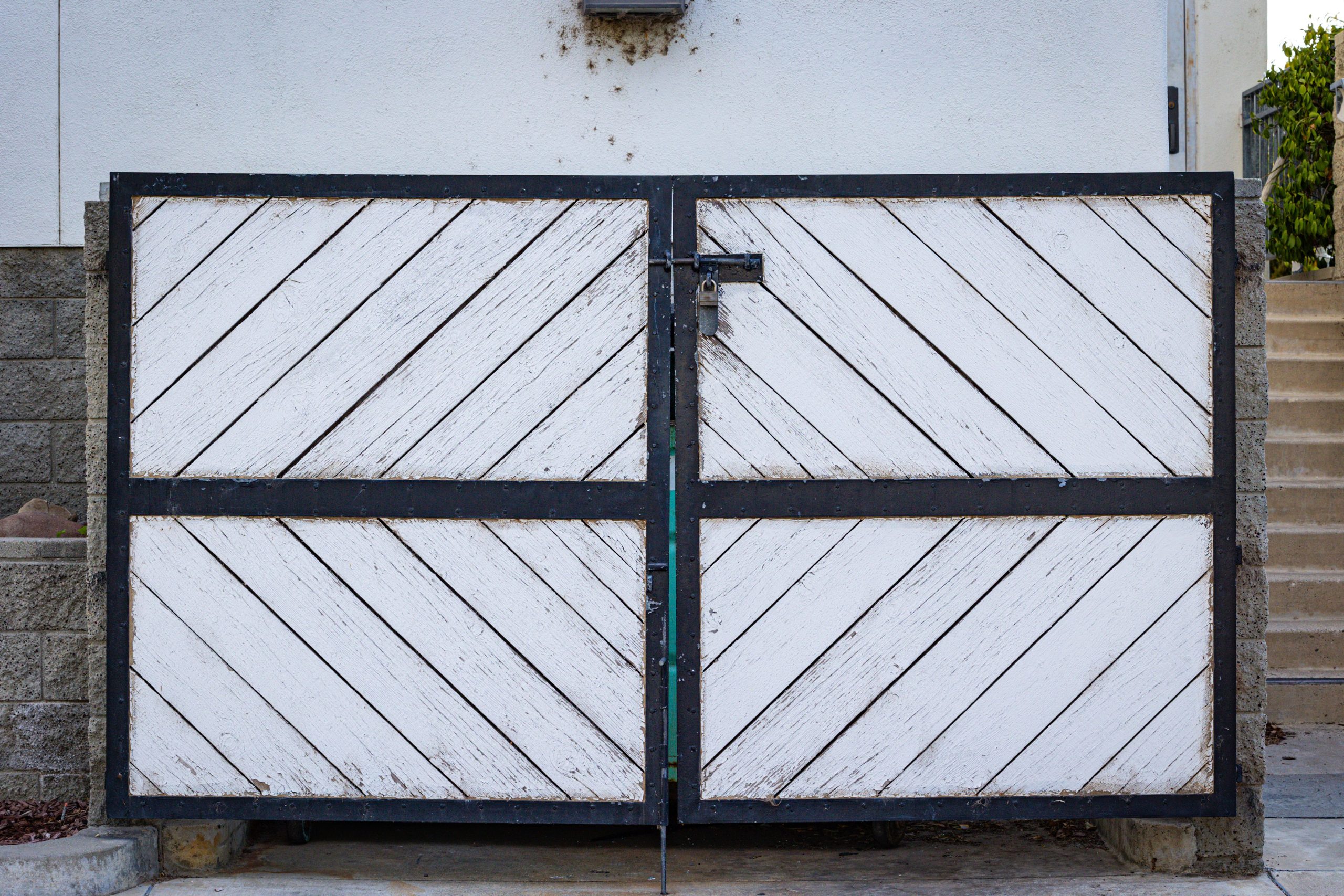 A rustic white wooden gate with diagonal panels and a black metal frame in a residential entrance. Un portail blanc rustique en bois avec des panneaux diagonaux et un cadre en métal noir à l'entrée d'une résidence.