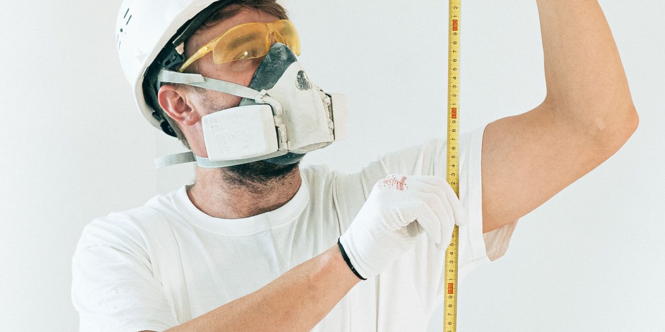 A construction worker in full safety gear measures with a tape against a white wall. Un ouvrier du bâtiment en équipement de sécurité complet mesure avec un mètre contre un mur blanc.