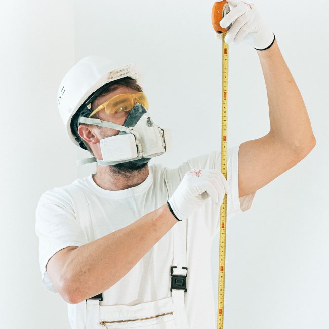 A construction worker in full safety gear measures with a tape against a white wall. Un ouvrier du bâtiment en équipement de sécurité complet mesure avec un mètre contre un mur blanc.