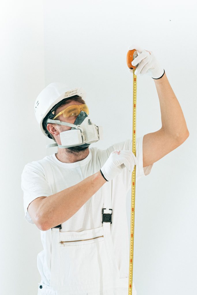 A construction worker in full safety gear measures with a tape against a white wall. Un ouvrier du bâtiment en équipement de sécurité complet mesure avec un mètre contre un mur blanc.