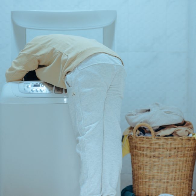 Back view full length barefoot faceless ethnic child in casual outfit standing on stool and taking clothes out of washing machine with top loader while keeping head inside of washer Vue arrière d'un enfant ethnique pieds nus sans visage en tenue décontractée debout sur un tabouret sortant des vêtements d'une machine à laver à chargement par le haut tout en gardant la tête à l'intérieur du lave-linge
