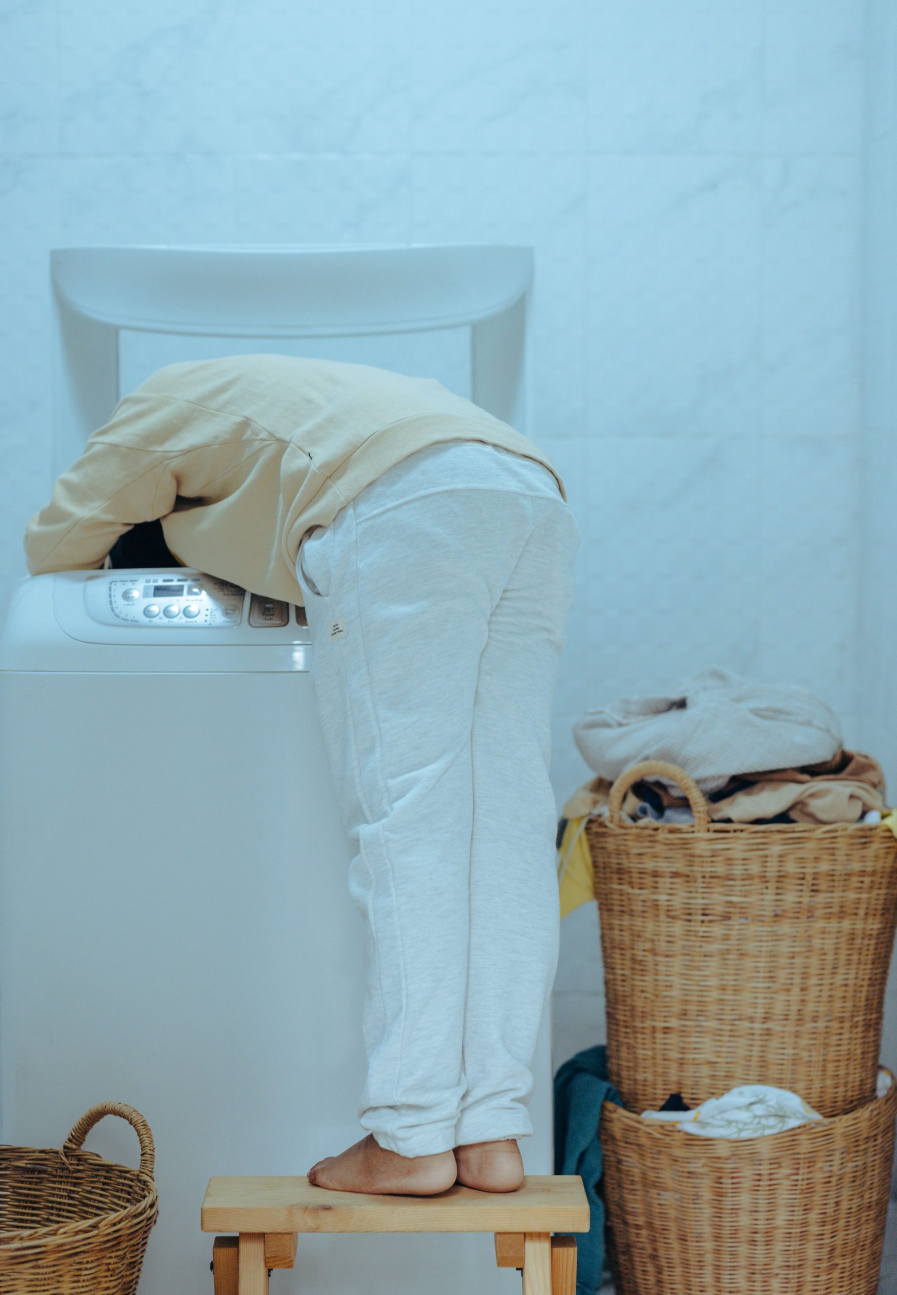 Back view full length barefoot faceless ethnic child in casual outfit standing on stool and taking clothes out of washing machine with top loader while keeping head inside of washer Vue arrière d'un enfant ethnique pieds nus sans visage en tenue décontractée debout sur un tabouret sortant des vêtements d'une machine à laver à chargement par le haut tout en gardant la tête à l'intérieur du lave-linge