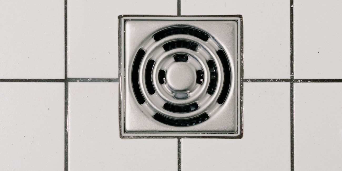 Top view of white ceramic tile floor with stainless steel square shaped cover of drain in contemporary shower Vue de dessus d'un sol en carrelage blanc avec grille d'évacuation carrée en acier inoxydable dans une douche contemporaine.