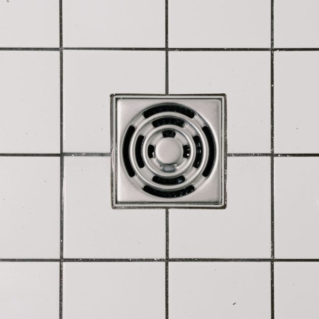 Top view of white ceramic tile floor with stainless steel square shaped cover of drain in contemporary shower Vue de dessus d'un sol en carrelage blanc avec grille d'évacuation carrée en acier inoxydable dans une douche contemporaine.