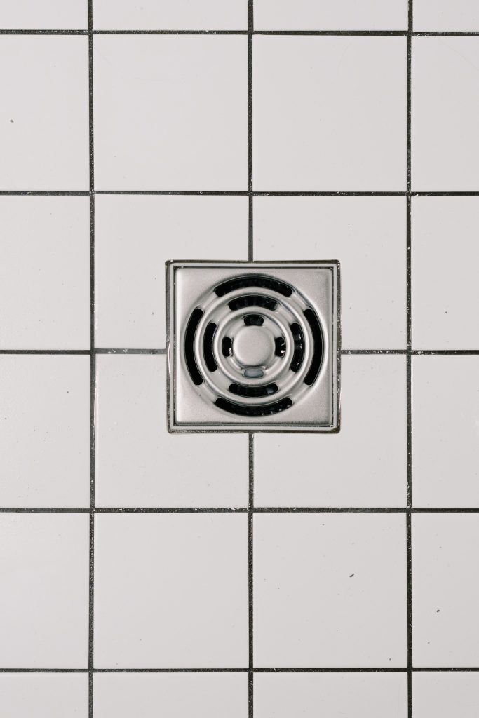 Top view of white ceramic tile floor with stainless steel square shaped cover of drain in contemporary shower Vue de dessus d'un sol en carrelage blanc avec grille d'évacuation carrée en acier inoxydable dans une douche contemporaine.